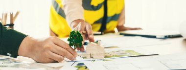 Profesyonel mimar ve mühendis, planlı, ahşap bloklu ve ağaç modelli bir toplantı masasında çevre dostu ev tasarlamak için işbirliği yapıyorlar. - Yakın çekim. Tanımlama.