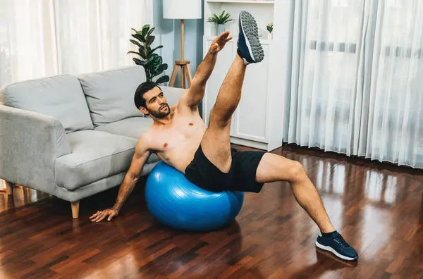 Athletic and sporty man doing situp on fitness ball during home body workout exercise session for fit physique and healthy sport lifestyle at home. Gaiety home exercise workout training concept.