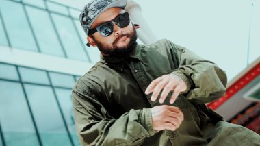 Asyalı hippi gökyüzü dövüşçüsüne dans ederken kameraya bakıyor. Çekici yakışıklı hippi ellerini alçak açılı kamerayla modern alışveriş merkezinde hip hop müziğine yöneltiyor. Açık hava sporu 2024. ÇalışmaComment.