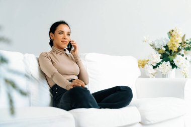 Rahat genç Afrikalı Amerikalı kadın evdeki oturma odasındaki koltukta çok önemli bir cep telefonu kullanıyor.