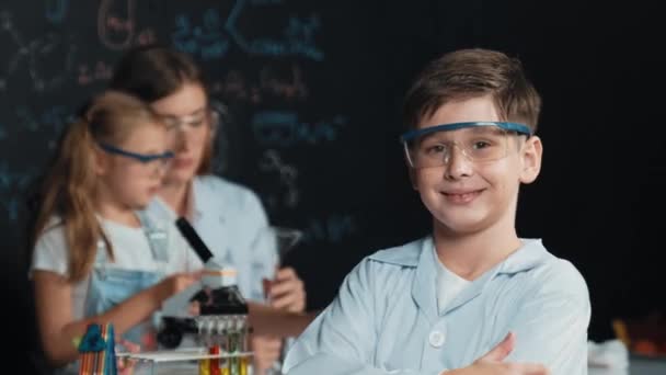 スマートな少年が腕を横断しながら 教師と友人がStemサイエンスクラスで実験をしています 学生は 実験室で化学理論を書かれたボードに立っている間 着色ソリューションを混合します エラー — ストック動画