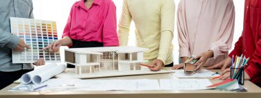 Profesyonel mimar takımının ev rengi seçimi hakkında beyin fırtınası yaparken, mimar da plan ve ev modelinin olduğu masadaki taslak belgeye yazı yazıyor. Değiştirilmiş.