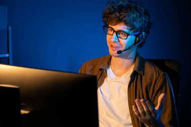 Genç yaratıcı iş adamı, bilgisayar ekranında dinleyicilerle iletişim kuruyor ya da takım üyesiyle görüşüyor. Neon Light Modern Ev Ofisi 'nde kulaklık takıyor. Gece vakti çalışma kavramı. Toplayıcı.