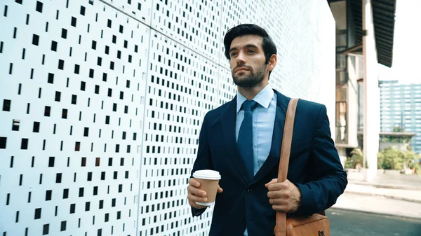Skilled business man walking in suit outfit while holding coffee cup. Professional manager standing near architectural building. Going to working in morning, seeking for successful job. Exultant.