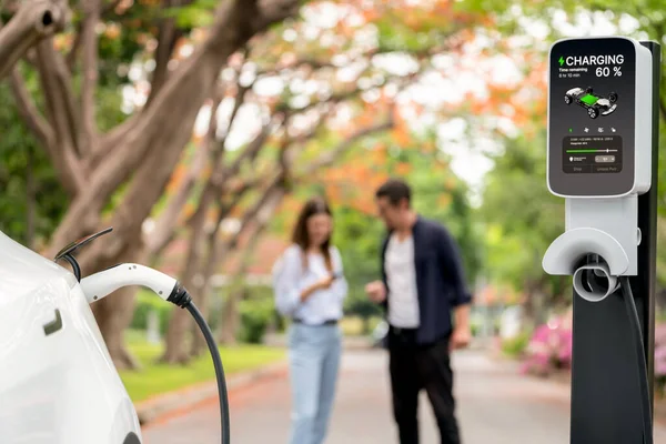 Focused EV car recharging battery on blurred background of lovey couple during autumnal road trip travel with electric vehicle recharging battery. Eco friendly travel on vacation during autumn. Exalt