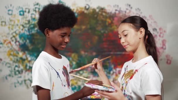 在艺术课上 在色彩斑斓的墙上 用油漆刷在彼此的白衬衫上涂上颜色 让不同的孩子们微笑 代表交流经验 互相学习 — 图库视频影像