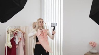 Genç sosyal medyum kadın selfie çubuğuyla moda videosu yapıyor. Blogcu kameraya gülümserken çevrimiçi kıyafetleri izleyicilere ya da takipçilere vlog sattırıyor. Blithe