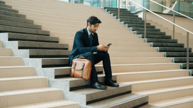 Akıllı proje yöneticisi merdivenlerde otururken cep telefonuna bakıyor. Çekici Kafkasyalı iş adamı telefon kullanarak çalışıyor. Profesyonel yatırımcı iş stratejisi planlamak için telefonu kullanıyor. Sevinçli..