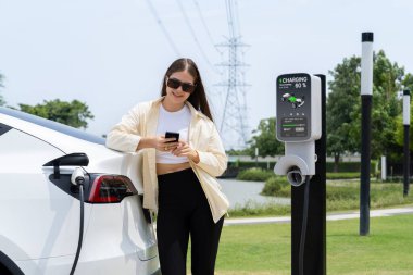 Genç bir kadın EV otomobil şarj istasyonu Green City Park 'ta elektrik ödemek için akıllı telefonu kullanıyor. Modern çevre ve EV araçlı sürdürülebilir şehir yaşam tarzı. Yararlı