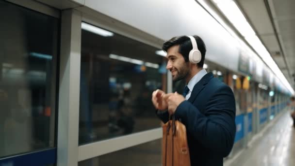 電車がプラットフォームで移動している間 ヘッドフォンから音楽を聞くスマートプロジェクトマネージャー ヘッドフォンを使用しているコーカサスビジネスマンは プラットフォームで待っている間リラックスした音楽を聞きます 仕事のルーチン エクササイズ — ストック動画