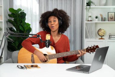Genç Afrikalı Amerikalı müzisyenin sunucu kanalı gitar çalıyor, şarkı söylüyor, stüdyoda dizüstü bilgisayardan yayın yapıyor. Kulaklık dekorasyonu ve mikrofon kaydı. Tatlandırıcı.