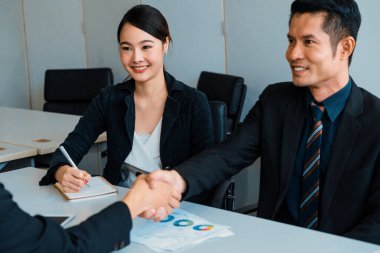 İş adamları konsepti kabul ediyor. Asyalı iş adamı ofis toplantı odasında başka bir iş adamıyla tokalaşıyor. Genç Asyalı sekreter onun yanında oturuyor. uds