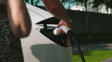 Genç bir kadın yazın yeşil sürdürülebilir şehir bahçesinde EV elektrikli arabasıyla seyahat ediyor. Kentsel sürdürülebilirlik yaşam tarzı yeşil temiz yeniden şarj edilebilir enerji elektrikli BEV araç iç organları