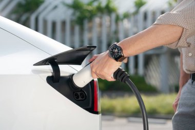 Genç adam, şehrin ticari otoparkındaki şarj istasyonundan elektrikli araba aküsünü şarj etmek için EV şarj aleti koymuş. Sürdürülebilir çevre dostu şehir yolculuğu için şarj edilebilir EV araba. Yararlı