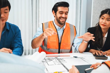 Mimari proje üzerinde birlikte çalışan çeşitli inşaat mühendisi ve müvekkil grupları, inşaat planını gözden geçirip toplantı masasında plan oluşturuyorlar. Tedbirli