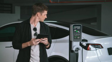 Genç adam EV elektrikli arabasıyla alışveriş merkezinin otoparkına gidiyor şehir merkezindeki şarj oluyor yeşil, temiz, yeniden şarj edilebilir elektrikli araç iç organları ile kentsel sürdürülebilirlik yaşam tarzını gösteriyor