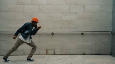 Asyalı akıllı hipster, kulaklık ve şık kıyafetler giyip hip hop şarkıları dinlerken gri arka planlı enerjik break dans gösterisi yapıyor. Açık hava sporu 2024. Hip hop hareketi. Yumuşak bir şekilde..