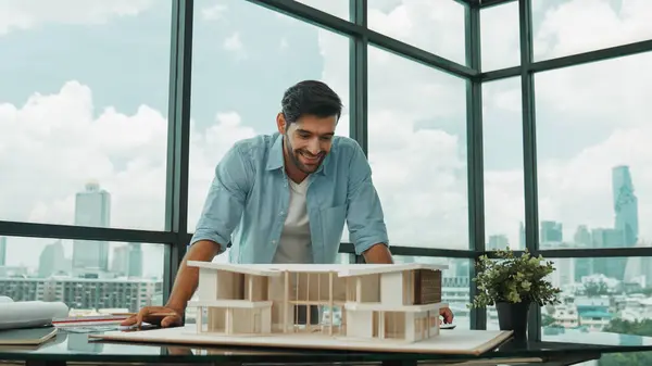 Portrait of engineer in casual outfit smiling at camera while inspect house model. Skilled architect looking at camera and standing near house model, project plan, architectural model. Tracery