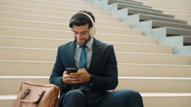 专业聪明的商人躺在楼梯上听音乐 聪明的经理听到好消息或从耳机里得到项目时很高兴 投资者坐着时穿着西服 膨胀剂 — 图库视频影像