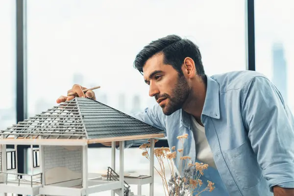 Attractive smart civil engineer measure house model roof construction. Professional architect in casual outfit looking at house model while planing building structure. Skyscraper view. Tracery.