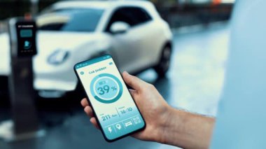 Akıllı telefon ekran bataryasını elinde tutan iş adamı akıllı EV mobil uygulama ile otomobil parkındaki şarj istasyonundan elektrik şarj eden çevre dostu EV otomobil. İnceleyin