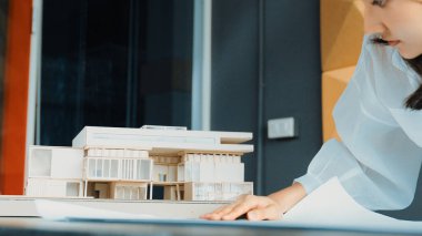 Asyalı genç ve güzel mimar mühendisimiz masaya model konulmuş proje planına bakıyor. Profesyonel proje yöneticisi ev yapısını analiz ediyor. İş tasarımı konsepti. Yönlendirici.