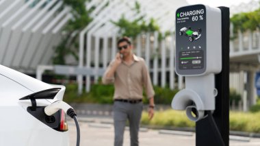 Genç adam telefonda konuşurken yeşil alışveriş merkezinin otoparkında EV elektrikli aracını şarj ediyor. Batarya şarj istasyonu olan çevre dostu EV arabası için sürdürülebilir kentsel yaşam tarzı. Panorama Çıkarı