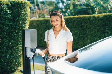 Genç bir kadın yazın yeşil sürdürülebilir şehir bahçesinde EV elektrikli arabasıyla seyahat ediyor. Kentsel sürdürülebilirlik yaşam tarzı yeşil temiz yeniden şarj edilebilir enerji elektrikli BEV araç iç organları