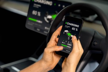 Aracın içindeki genç kadın, modern EV arabasındaki akıllı telefon ekranında akü şarj etme uygulamasını kontrol ediyor. Yükselt