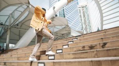 Asyalı şık bir adam alçak açılı kamerayla hip hop dansı yapıyor. Break dansçı, şehirde kulaklıktan müzik dinleyip dans ederken merdivenlerden çıkıyor. Açık hava sporu 2024. Yumuşak bir şekilde..