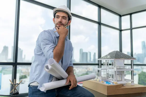 Portrait of professional architect engineer thinking about modern house project while wearing safety helmet and casual outfit. Smart interior designer holding project plans near house model. Tracery.