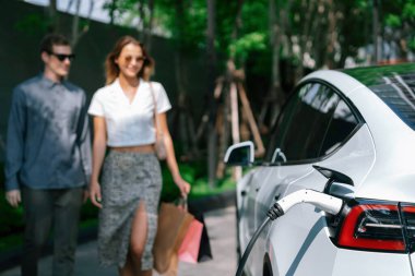 Genç çift yazın yeşil sürdürülebilir şehir bahçesinde EV elektrikli araba ile seyahat ederken, yeşil temiz yeniden şarj edilebilir elektrikli araç iç organları ile kentsel sürdürülebilirlik yaşam tarzını gösteriyor.