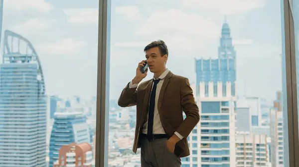 Businessman standing in ornamented office talking on phone with college on cityscape skyline window background. Determination and ambition drive business career toward to bright future