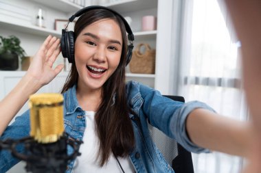 Sunucu kanalı genç ve güzel Asyalı kız akıllı telefonuyla selfie çekiyor. Modern stüdyo ofis konseptinde kulaklık takıyor. Sahne arkası yayıncısı sosyal medyada canlı yayın yapıyor. Taktik.