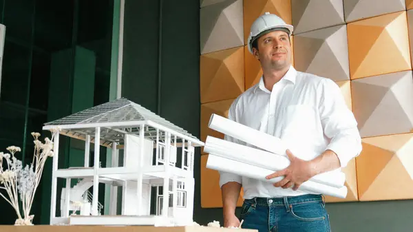 Portrait of professional project manager holding project plan with house model at table. Professional architect engineer wearing safety helmet while standing posing with confident. Manipulator.