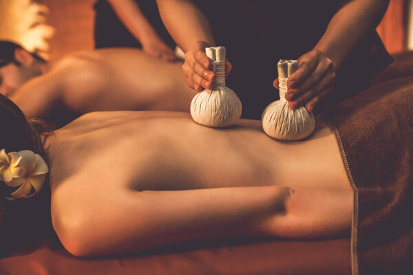 Hot herbal ball spa massage body treatment, masseur gently compresses herb bag on couple customer body. Serenity of aromatherapy recreation in warm lighting of candles at spa salon. Quiescent