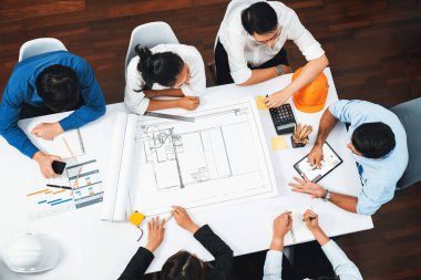 Mimari proje üzerinde birlikte çalışan, inşaat planını gözden geçiren ve toplantı masasında plan oluşturan çeşitli inşaat mühendisi ve müşteri gruplarından oluşan üst düzey bir pankart. Tedbirli