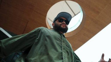 Kameraya bakarken Asyalı yakışıklı hipsterin hip hop pozu. Profesyonel sokak dansçısının alışveriş merkezinde serbest dans edişinin düşük açılı görüntüsü. Açık hava sporu 2024. ÇalışmaComment.
