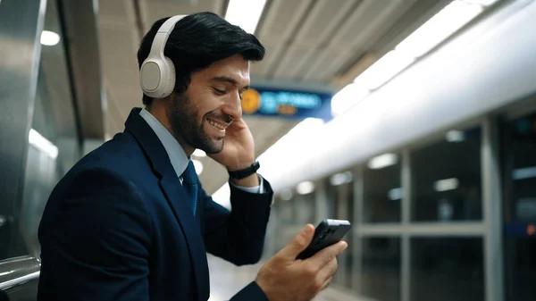 Smart business man listening music by headphone while waiting train at train station with blurring background. Skilled project manager enjoy listen relax sound while holding mobile phone. Exultant.