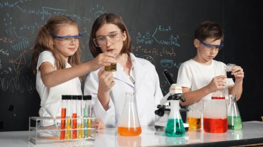 Öğretmen tavsiye verirken beyaz çocuk kimyasal sıvıyı karıştırıyor. Profesyonel eğitmen labaratuar kıyafeti giymiş, renkli solüsyonla dolu deney kabıyla sofrada çeşitli öğrencileri arıyor. Etkinlik.