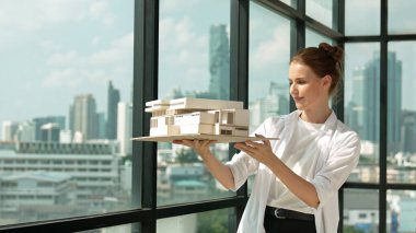 Genç, beyaz, güzel mimar mühendisi ev modelini yükseltirken şehir gökdelen manzarası dikkatlice inceleniyor. İnşaat mühendisliği, mimari stüdyosu. Proje planlama. İzleme