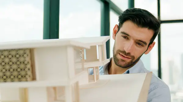 Skilled smiling caucasian engineer turns house model left and right to check mistake point. Professional manager inspect, checking, looking at architectural model. Design concept. Tracery