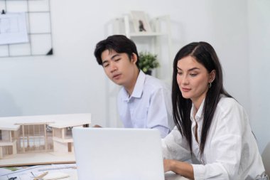 Profesyonel Asyalı erkek mimar ev modelinin uzunluğunu ölçmek için cetvel kullanırken, beyaz tenli genç meslektaşı ev modeliyle toplantı masasındaki verileri analiz etmek için dizüstü bilgisayar kullanıyor. Tertemiz..