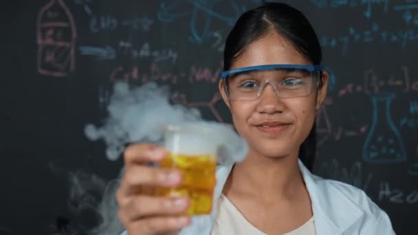 スマートな高校生のクローズアップは ブラックボードで着色されたソリューションを検査します ビーカーを持ち ラボの黒板の前に立っている間化学液体を見ている美しい科学者 イスラーム主義 — ストック動画