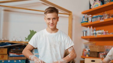 Çeşitli çocuklar sanat dersinde çalışırken mutlu genç modellik kupası ve kameraya bakmak. Çeşitli lise öğrencileri atölyede yaratıcı faaliyetler yaparken ayakta duruyorlar. Öğretim.