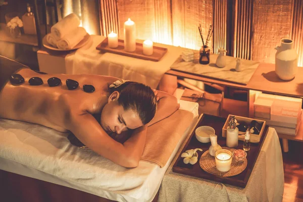 Hot stone massage at spa salon in luxury resort with warm candle light, blissful woman customer enjoying spa basalt stone massage glide over body with soothing warmth. Quiescent