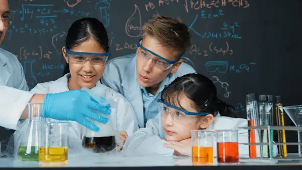 Girl looking at beaker while doing experiment at blackboard with chemistry theory in STEM class. teacher and group of highschool student with mixed races doing science activity together. Edification.
