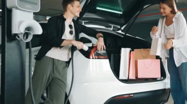 Yavaş çekim çift EV elektrikli arabayla şehir merkezindeki alışveriş merkezine gidiyor. Şehir merkezinde yeşil, temiz, yeniden şarj edilebilir elektrikli araç iç organları tarafından sürdürülebilirlik yaşam biçimi olarak şarj ediliyor.