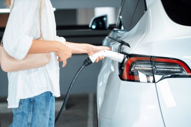 Genç bir kadın EV elektrikli arabasıyla alışveriş merkezinin otoparkına gidiyor şehir merkezinde elektrik iç organlarının yeşil temiz şarj edilebilir enerji ile sürdürülebilirlik yaşam tarzını gösteriyor.
