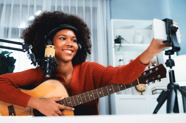 Akıllı telefon ayarlayan güzel Afrikalı kadın nüfuzlu sunucu kanalı yayında gitarla şarkı söylüyor. Canlı sosyal medyada müzik blogcusu zamanı. Ses yaratıcısı kavramı. Tatlandırıcı.
